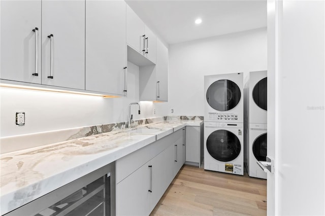 clothes washing area with beverage cooler, cabinet space, a sink, stacked washer and clothes dryer, and light wood-type flooring