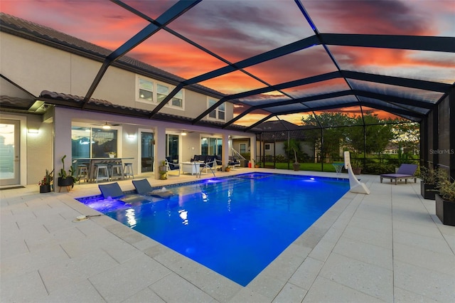 pool at dusk featuring glass enclosure, a patio area, and a bar