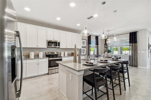 kitchen with stainless steel appliances, light stone counters, an island with sink, pendant lighting, and white cabinets