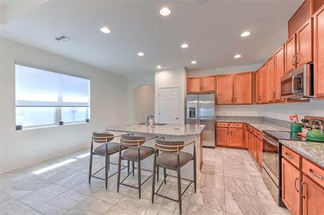 kitchen with a breakfast bar, light stone counters, an island with sink, and appliances with stainless steel finishes