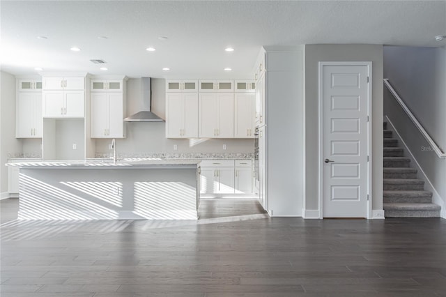 kitchen with dark wood-type flooring, visible vents, white cabinetry, wall chimney range hood, and an island with sink