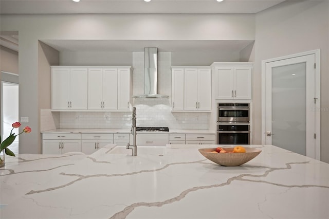 kitchen with white cabinets, wall chimney exhaust hood, light stone countertops, and stainless steel appliances