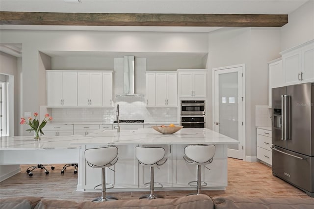 kitchen featuring appliances with stainless steel finishes, light stone counters, wall chimney range hood, beamed ceiling, and white cabinetry