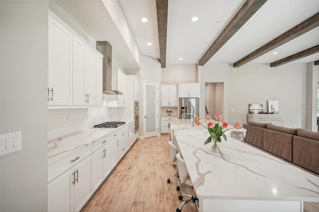 kitchen featuring beam ceiling, white cabinetry, wall chimney exhaust hood, a spacious island, and appliances with stainless steel finishes