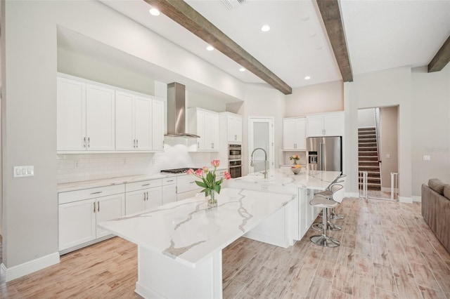 kitchen featuring a large island with sink, white cabinets, stainless steel appliances, and wall chimney range hood