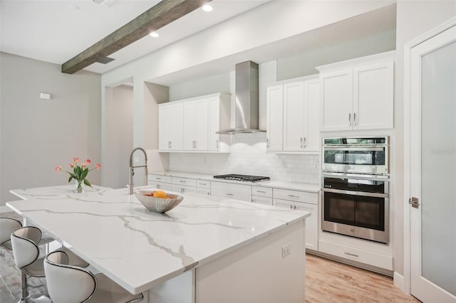 kitchen with beam ceiling, a kitchen island with sink, white cabinets, and wall chimney range hood