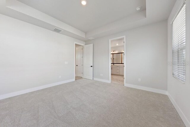 unfurnished bedroom featuring a raised ceiling, ensuite bathroom, and light colored carpet