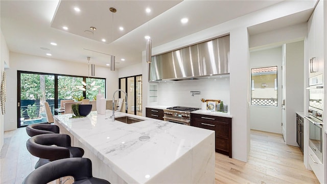 kitchen with light stone countertops, stainless steel appliances, wall chimney range hood, pendant lighting, and a center island with sink