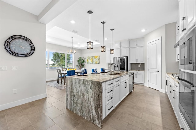 kitchen featuring light stone counters, stainless steel appliances, decorative light fixtures, a center island with sink, and white cabinets