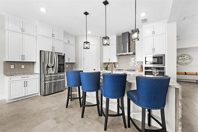 kitchen featuring white cabinetry, wall chimney exhaust hood, light stone counters, a kitchen island with sink, and appliances with stainless steel finishes