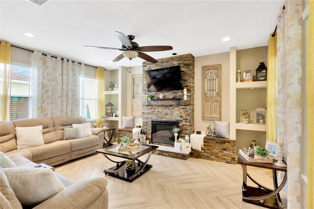 living room with built in shelves, ceiling fan, a stone fireplace, and light parquet flooring