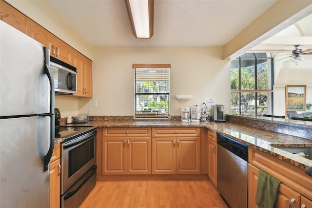 kitchen featuring kitchen peninsula, appliances with stainless steel finishes, light wood-type flooring, ceiling fan, and dark stone countertops