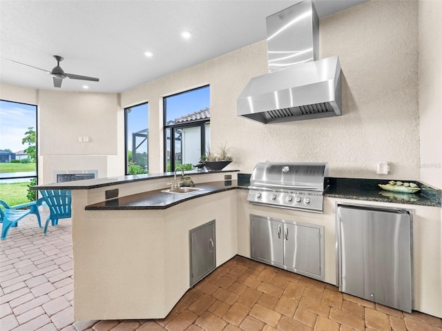 kitchen with dark countertops, refrigerator, a peninsula, wall chimney range hood, and a sink