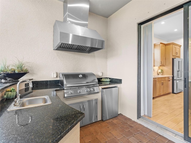 kitchen featuring high end refrigerator, dark countertops, freestanding refrigerator, wall chimney range hood, and a sink