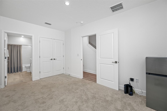 unfurnished bedroom featuring ensuite bathroom, a textured ceiling, light colored carpet, stainless steel refrigerator, and a closet