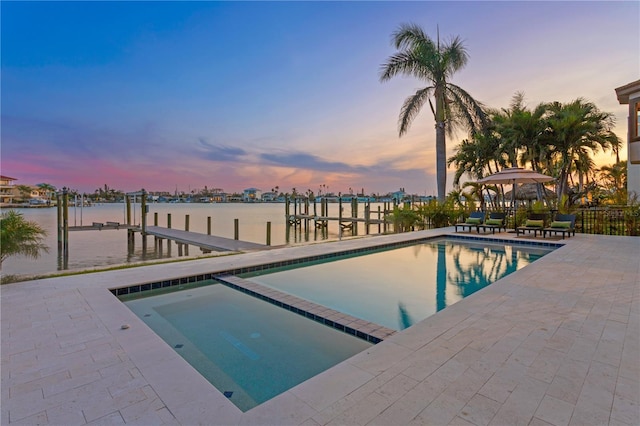 pool at dusk with a pool with connected hot tub, a water view, a boat dock, and a patio