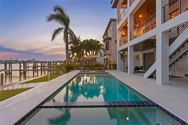 pool at dusk featuring a ceiling fan, a pool with connected hot tub, a water view, and a patio