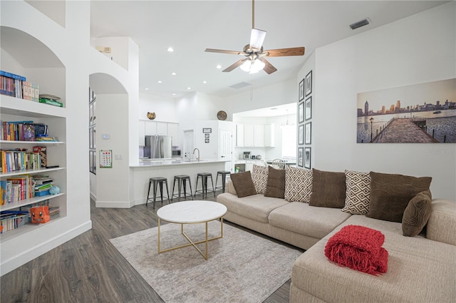 living room with built in shelves, ceiling fan, sink, hardwood / wood-style floors, and a towering ceiling