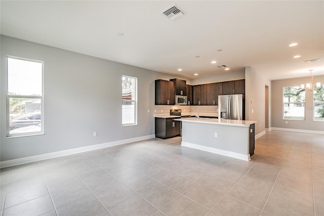 kitchen featuring stainless steel appliances, an island with sink, an inviting chandelier, light tile patterned flooring, and dark brown cabinets