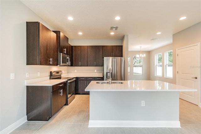 kitchen featuring stainless steel appliances, an inviting chandelier, light tile patterned floors, an island with sink, and dark brown cabinets