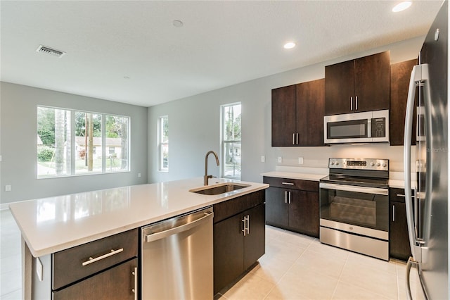 kitchen with appliances with stainless steel finishes, an island with sink, light tile patterned floors, dark brown cabinetry, and sink