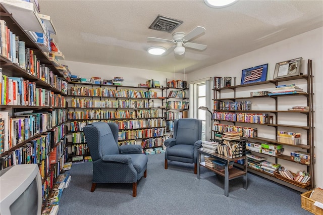 sitting room featuring visible vents, a ceiling fan, carpet flooring, wall of books, and a textured ceiling