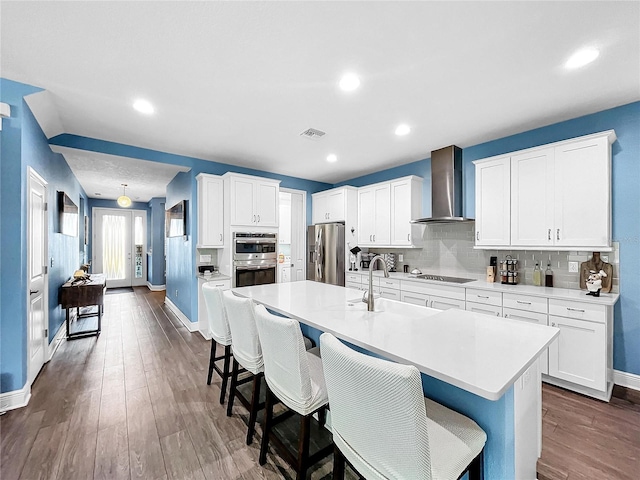 kitchen featuring white cabinets, an island with sink, wall chimney range hood, and appliances with stainless steel finishes