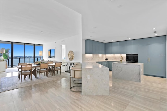 kitchen with light stone counters, a wall of windows, a sink, decorative backsplash, and an island with sink