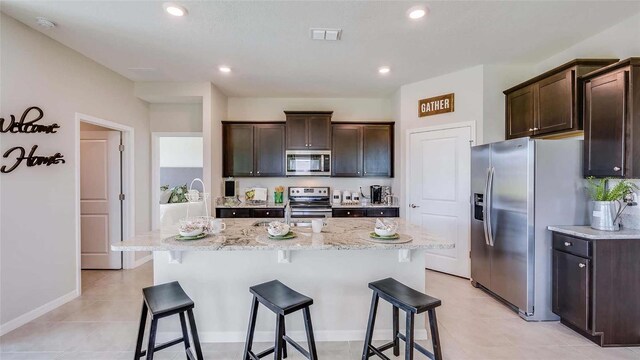 kitchen featuring stainless steel appliances, an island with sink, light stone counters, dark brown cabinetry, and light tile floors