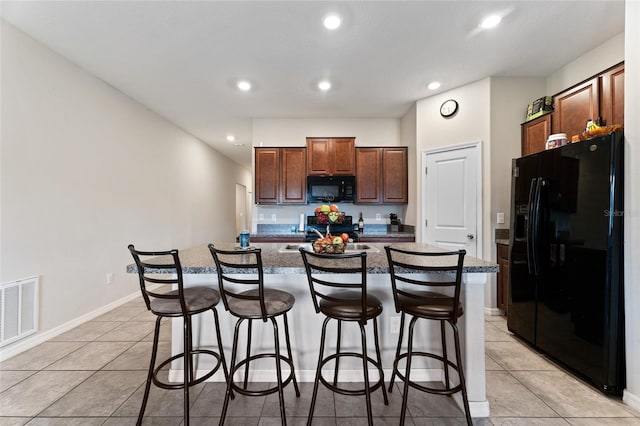 kitchen featuring an island with sink, black appliances, a breakfast bar area, stone countertops, and light tile floors