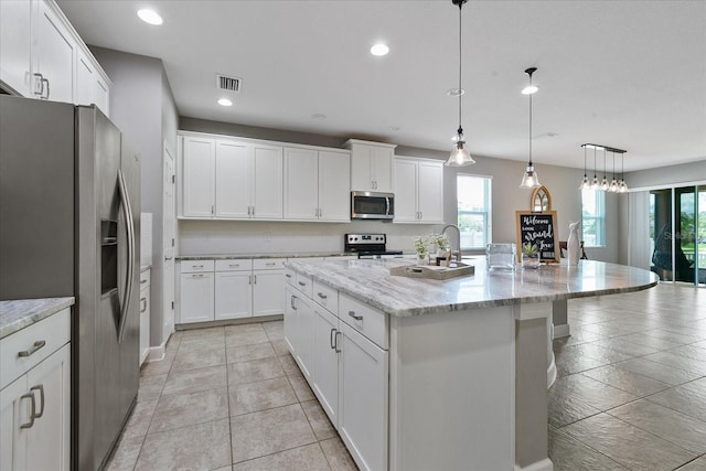 kitchen with white cabinets, hanging light fixtures, an island with sink, light stone counters, and stainless steel appliances