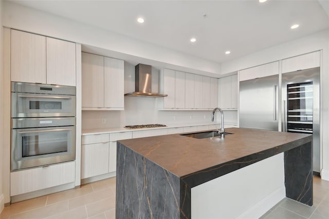 kitchen with stainless steel appliances, sink, wall chimney range hood, light tile patterned floors, and a center island with sink