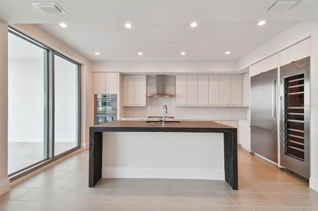 kitchen featuring stainless steel appliances, wall chimney range hood, white cabinets, plenty of natural light, and an island with sink