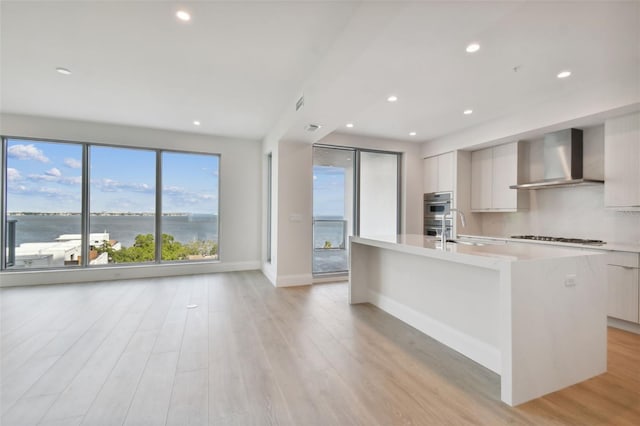 kitchen featuring wall chimney exhaust hood, sink, a water view, a center island with sink, and white cabinetry