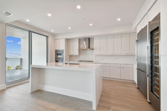 kitchen featuring white cabinetry, a center island with sink, and wall chimney range hood