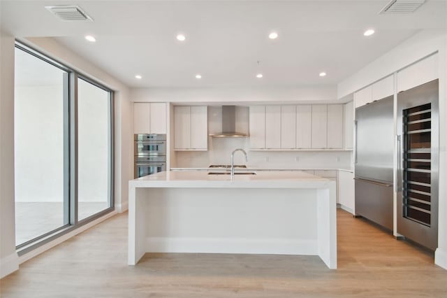 kitchen with white cabinets, light wood-type flooring, wall chimney range hood, and an island with sink