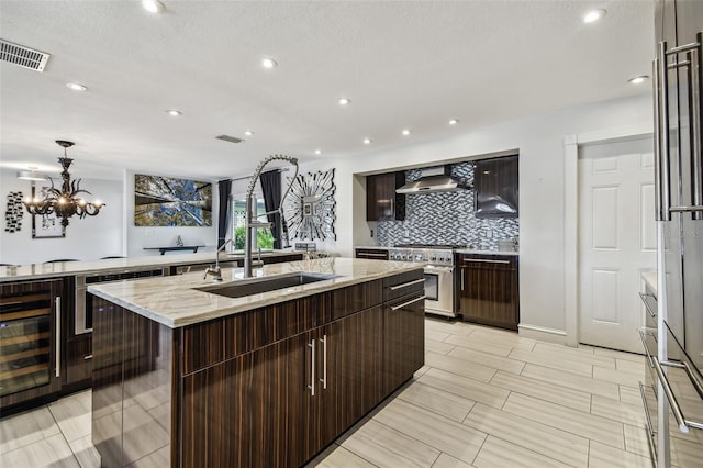 kitchen featuring wall chimney exhaust hood, sink, an island with sink, pendant lighting, and high end stove