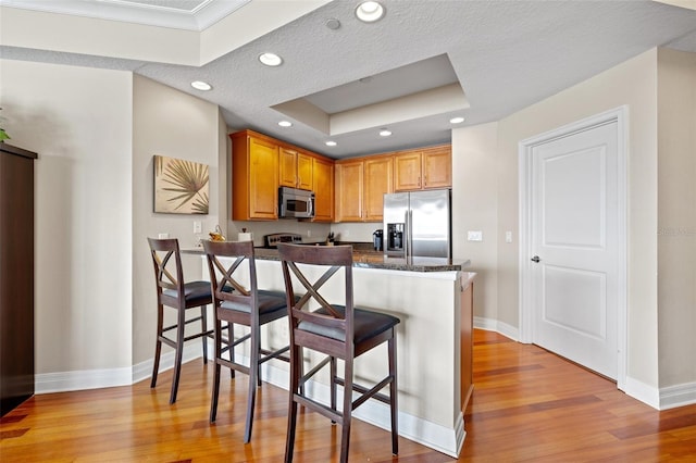 kitchen featuring stainless steel appliances, kitchen peninsula, dark stone counters, a tray ceiling, and a kitchen bar