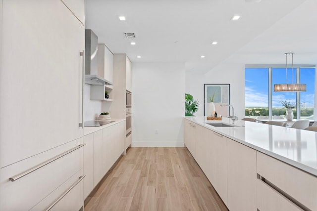 kitchen featuring sink, wall chimney exhaust hood, decorative light fixtures, light wood-type flooring, and white cabinetry