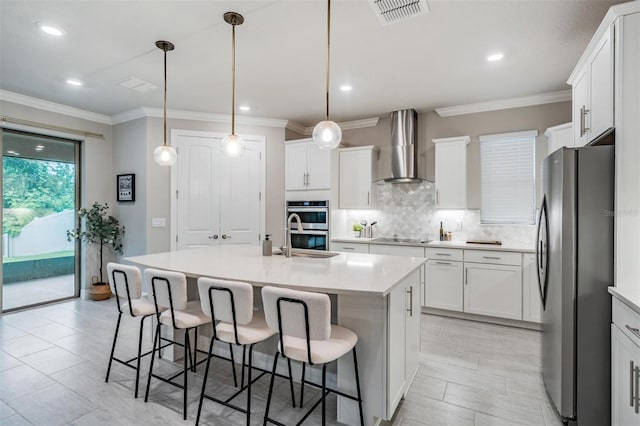 kitchen with appliances with stainless steel finishes, wall chimney range hood, an island with sink, decorative backsplash, and hanging light fixtures