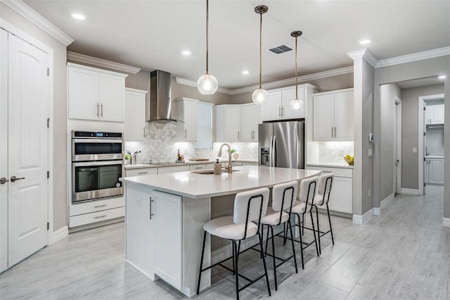 kitchen with stainless steel appliances, an island with sink, white cabinets, and wall chimney range hood