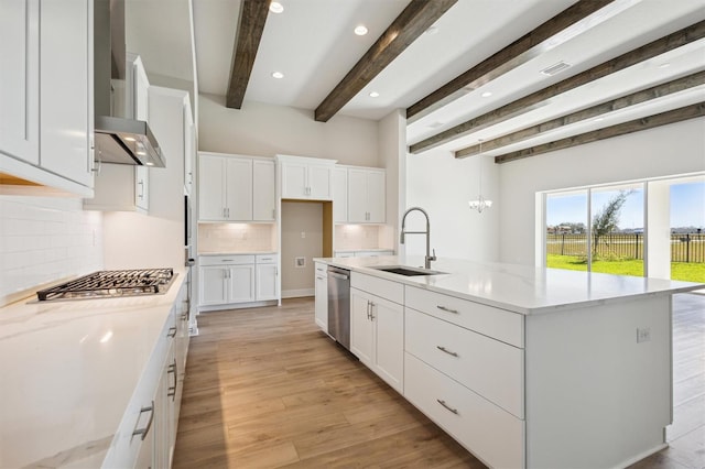 kitchen with wall chimney range hood, beamed ceiling, a center island with sink, appliances with stainless steel finishes, and white cabinets