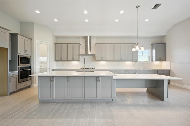 kitchen featuring stainless steel appliances, a kitchen island with sink, wall chimney range hood, decorative light fixtures, and gray cabinets