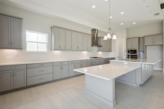 kitchen with gray cabinetry, gas stovetop, wall chimney range hood, decorative light fixtures, and an island with sink