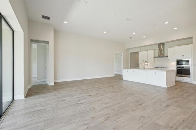 kitchen with white cabinets, light hardwood / wood-style flooring, stainless steel appliances, wall chimney exhaust hood, and a kitchen island with sink