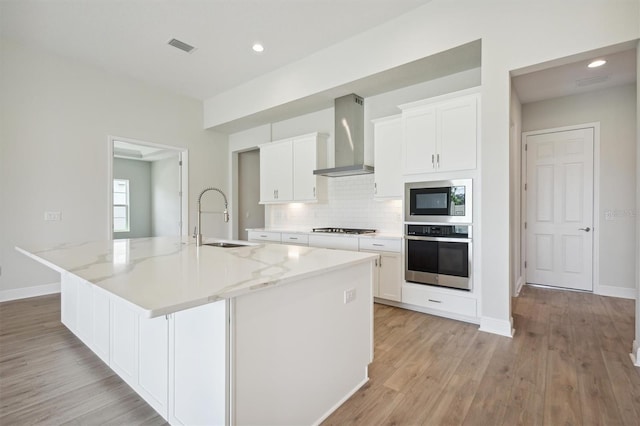 kitchen with wall chimney exhaust hood, a center island with sink, appliances with stainless steel finishes, sink, and light wood-type flooring