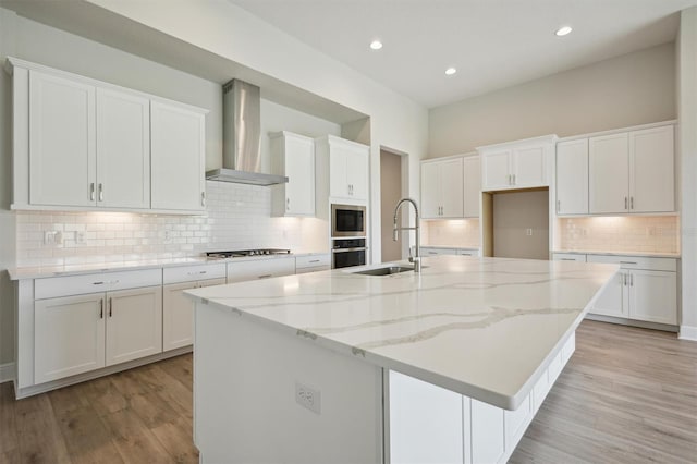 kitchen with wall chimney exhaust hood, light hardwood / wood-style flooring, a kitchen island with sink, sink, and white cabinets