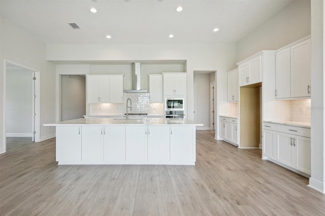 kitchen with light hardwood / wood-style flooring, white cabinetry, wall chimney range hood, stainless steel oven, and a kitchen island with sink