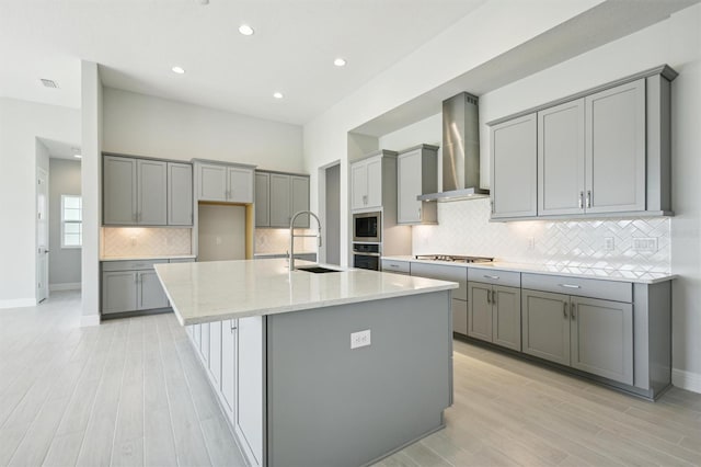 kitchen featuring wall chimney range hood, a center island with sink, sink, gray cabinets, and appliances with stainless steel finishes