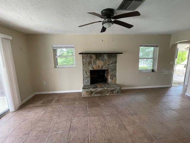 unfurnished living room with a textured ceiling, ceiling fan, a stone fireplace, and tile patterned floors